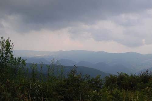 A panoramic view of rolling mountains under a cloudy sky, with lush greenery in the foreground.