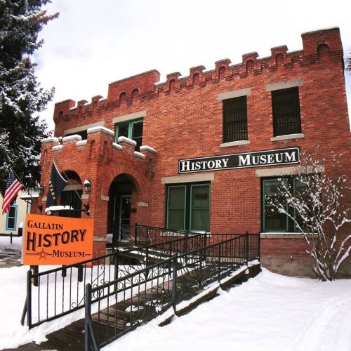 Historic brick building with a castle-like design, featuring a sign for the Gallatin History Museum and snow-covered surroundings.