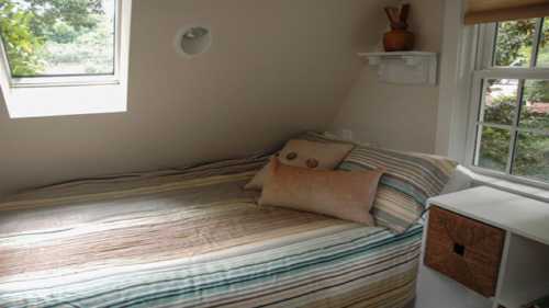 Cozy bedroom with a sloped ceiling, featuring a striped bedspread, pillows, and a small shelf by the window.