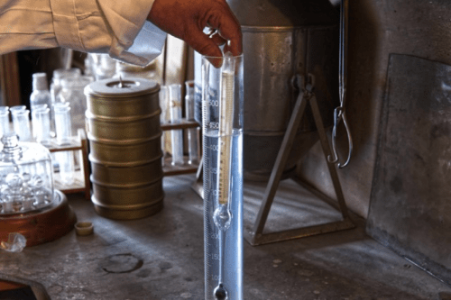 A hand holds a graduated cylinder filled with liquid, surrounded by various laboratory glassware and equipment.