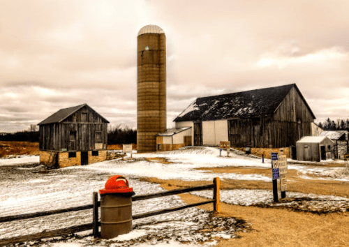 A rustic farm scene featuring a barn, silo, and snow-covered ground under a cloudy sky.