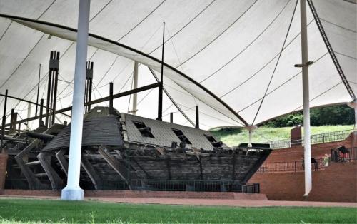 A historic shipwreck displayed under a large white canopy, surrounded by green grass and brick structures.