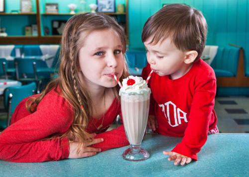 A girl and a boy share a milkshake with whipped cream and a cherry, both smiling and sipping through straws.