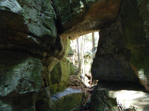 A narrow passage between large moss-covered rocks, with trees visible in the background. Natural light filters through.