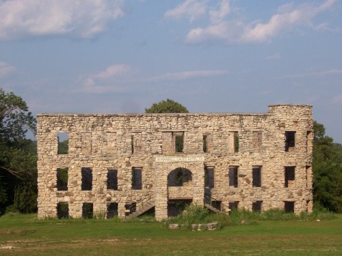 Ruins of a stone building with large windows, set against a blue sky and green landscape.