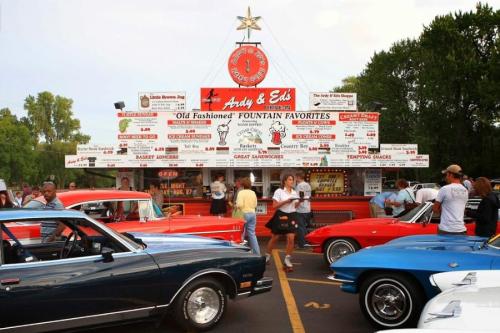 A busy drive-in diner with a large menu sign, surrounded by classic cars and people enjoying the atmosphere.