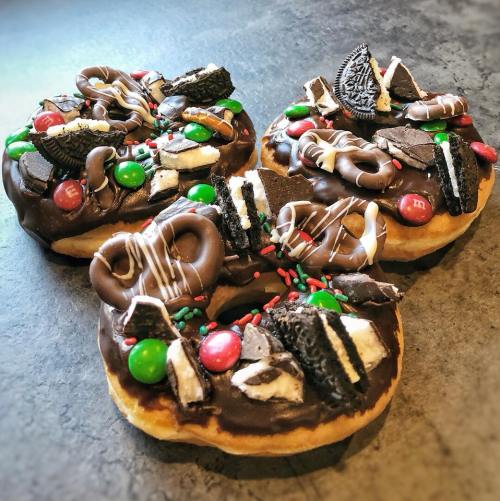 Three chocolate-covered donuts topped with pretzels, Oreos, M&M's, and festive sprinkles.