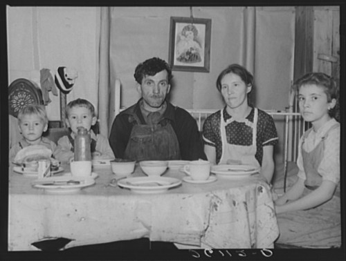 A family of five sits at a table with empty dishes, in a modest home setting. Two children are on the left.