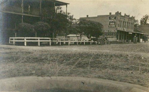 Black and white photo of a small town street featuring a drug store and nearby buildings with a dirt road in the foreground.