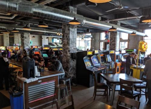 A lively arcade and bar with vintage game machines, patrons enjoying drinks, and a modern industrial decor.