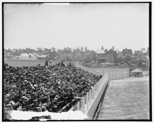 A large crowd in hats fills a stadium, with houses visible in the background on a sunny day.