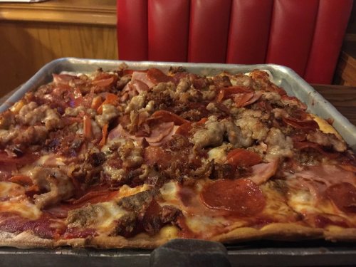 A close-up of a large, loaded pizza topped with various meats, including pepperoni, sausage, and ham, on a metal tray.