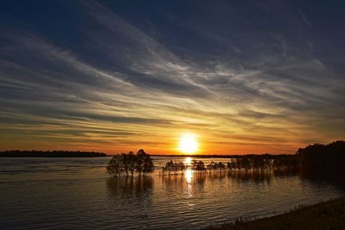 A serene sunset over a calm river, with silhouetted trees and colorful clouds reflecting on the water's surface.