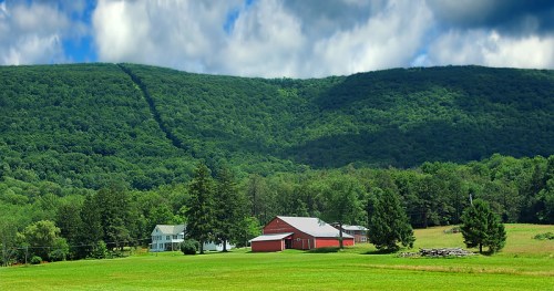 A scenic view of a green hillside with a red barn and farmhouse in a lush, rural landscape under a partly cloudy sky.