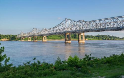 A steel bridge spans a river, surrounded by green hills and blue skies, reflecting the sunlight on the water.