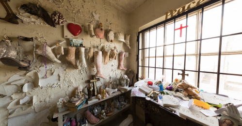 A cluttered room with various objects, including decorated boots, figurines, and a cross, against a peeling wall.