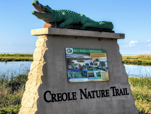 Sign for Creole Nature Trail featuring a green alligator sculpture and information about the area's ecology.