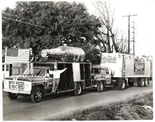 A vintage black-and-white photo of two trucks, one with a large soda bottle, promoting "Barq's Root Beer."