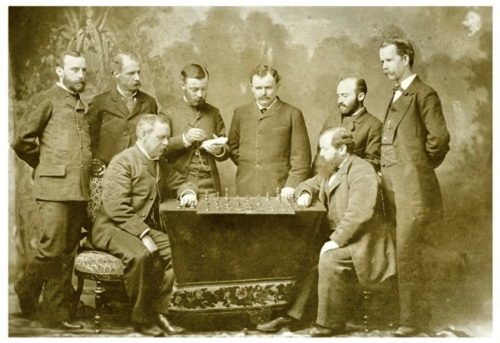 A historic black-and-white photo of eight men gathered around a chessboard, engaged in a game.
