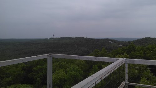 View from a lookout tower overlooking a dense green forest and distant hills under a cloudy sky.