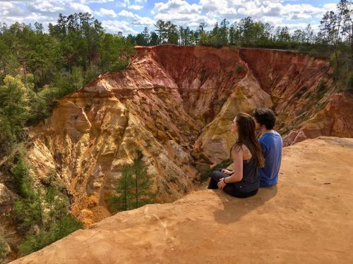 A couple sits on a cliff edge, overlooking a colorful canyon surrounded by trees and a blue sky with clouds.