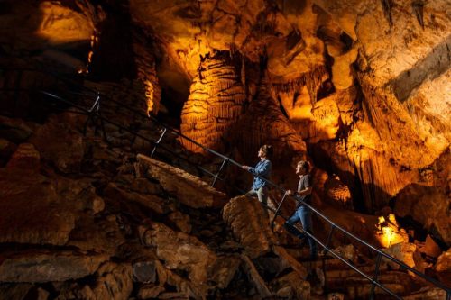 Two people ascend a metal staircase in a dimly lit cave, surrounded by rock formations and warm lighting.