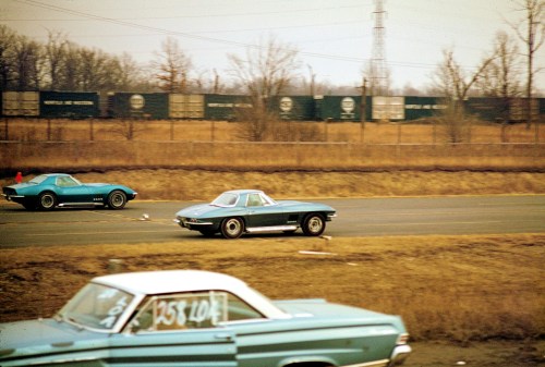 Two classic cars racing on a highway, with a blurred background of trees and train cars in the distance.