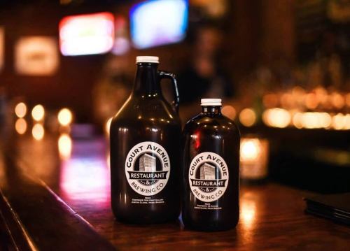 Two dark glass growlers labeled "Court Avenue Restaurant & Brewing Co." sit on a wooden bar. Soft lighting in the background.