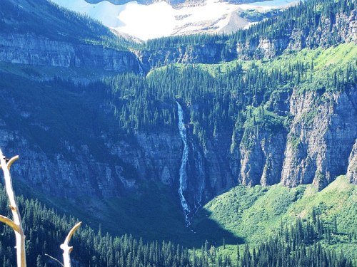 A scenic view of a waterfall cascading down a rocky cliff surrounded by lush green forests and mountains.