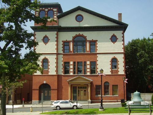Historic brick building with large windows and decorative elements, surrounded by trees and a clear blue sky.