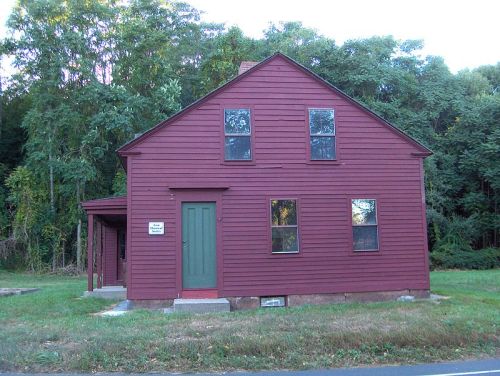 A red wooden house with green door and windows, surrounded by trees and grass.