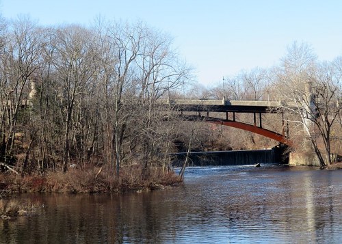A bridge arches over a calm river, surrounded by bare trees and a clear blue sky.