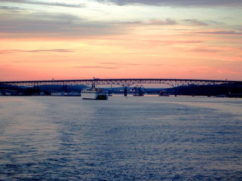 A serene sunset over a river, featuring a bridge and a boat gliding through calm waters.