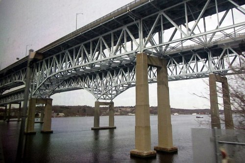 A steel bridge spans over a calm river, supported by tall concrete pillars, with boats visible in the background.