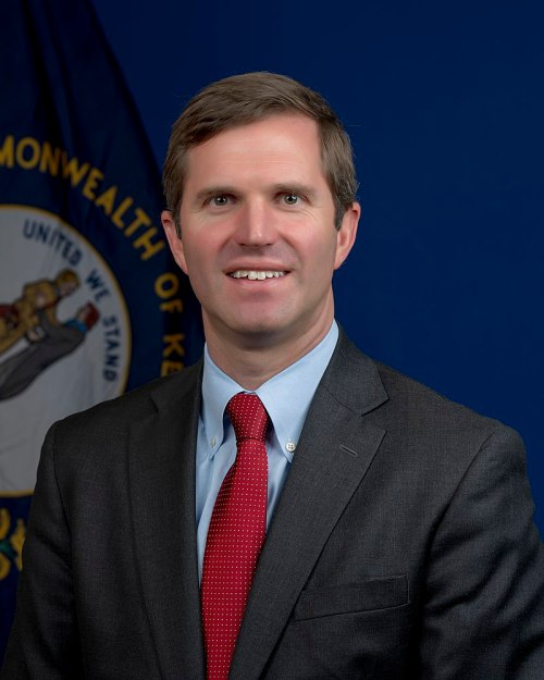A man in a suit and red tie smiles in front of a blue background with a state flag.