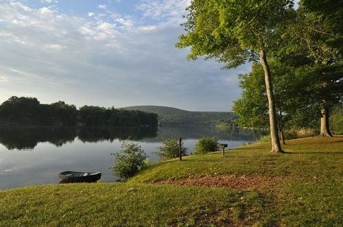 A serene lake scene with trees, a grassy shore, and gentle hills under a cloudy sky. A small boat is visible on the water.