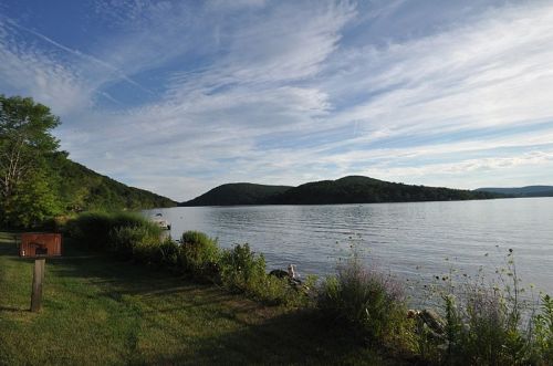 A serene lakeside view with lush greenery, rolling hills, and a clear blue sky reflected in the calm water.