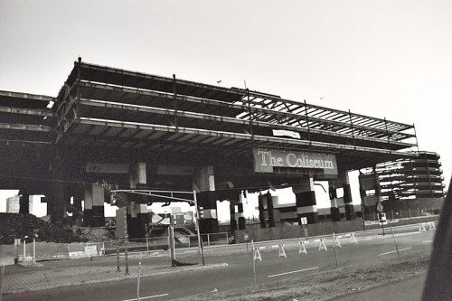 Black and white image of a partially constructed building labeled "The Coliseum," with surrounding barriers and debris.
