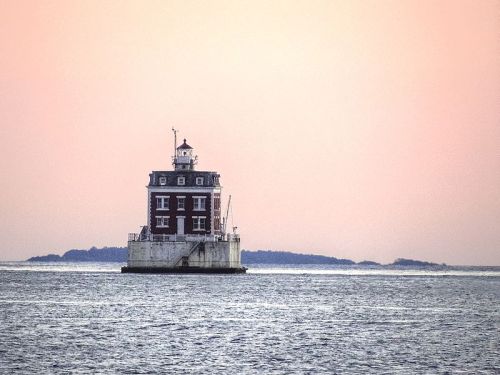 A historic lighthouse stands on a rocky base in the water, with a soft pastel sky in the background.