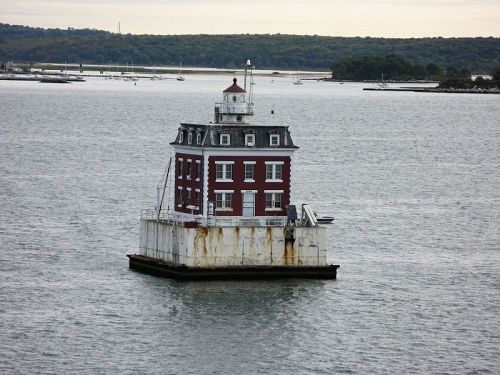 A red and white lighthouse stands on a small platform in the water, surrounded by calm waves and distant land.