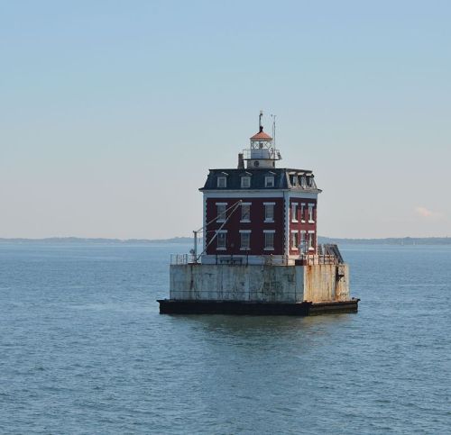 A red lighthouse stands on a rocky base in the middle of calm blue waters under a clear sky.