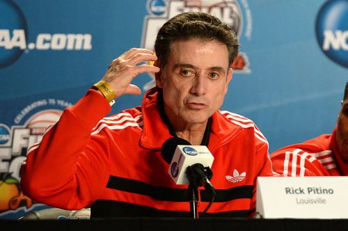 Rick Pitino speaking at a press conference, wearing a red Adidas jacket, gesturing with his hand.