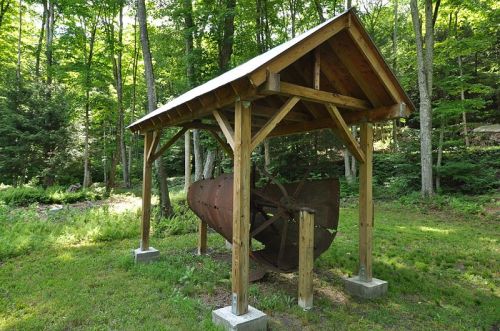 A wooden shelter in a forest houses a large, rusty metal drum on a concrete base, surrounded by greenery.