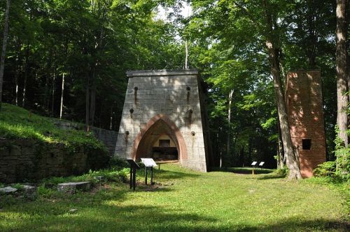 Historic stone structure surrounded by trees, with informational signs and a brick chimney nearby.