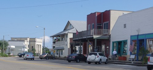 A small town street lined with historic buildings, shops, and parked cars under a clear blue sky.