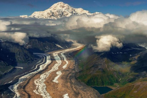 Aerial view of a glacier winding through mountains, with a rainbow and clouds surrounding a snow-capped peak.