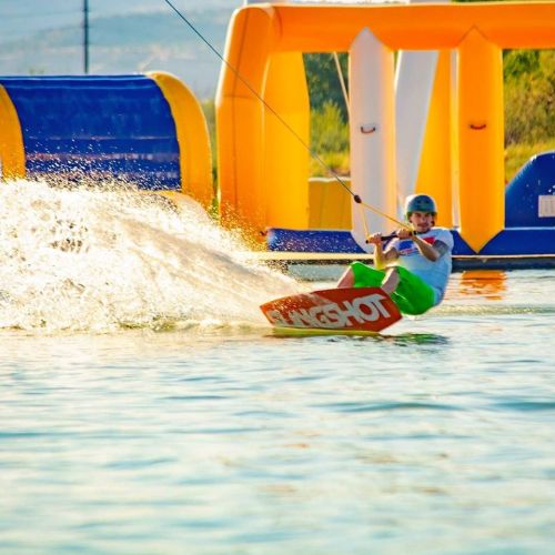 A person wakeskating on water, splashing as they maneuver near inflatable obstacles in a sunny setting.