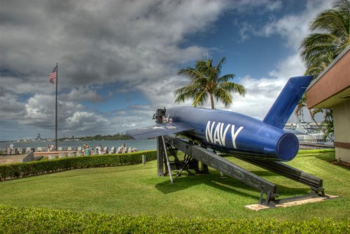 A blue Navy missile displayed on a stand, with palm trees and a cloudy sky in the background near a waterfront.