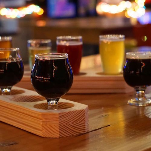 A wooden flight tray holds various small glasses of colorful craft beers on a bar table, with blurred lights in the background.
