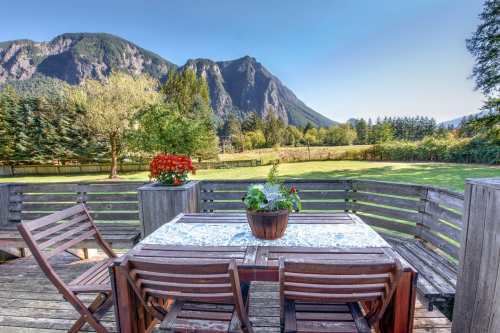 A wooden deck with a table and chairs overlooks a scenic landscape of mountains and greenery under a clear blue sky.